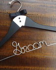 A luxe matte black hanger, hand-painted with a tuxedo design and finished with two tiny buttons and a grosgrain bowtie, customized with "Groom," in silver wire with notches. On a wooden background.