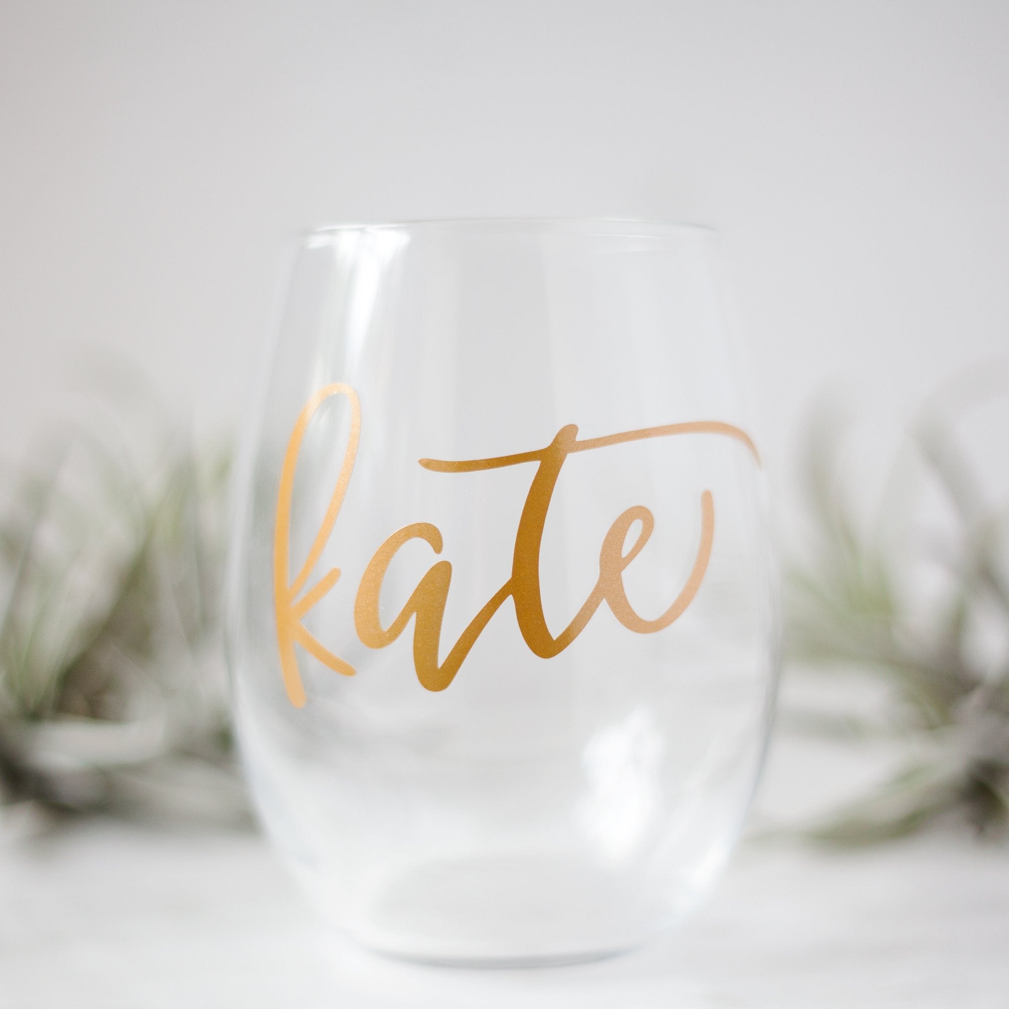 Stemless Wine Glass, shown in the image, is a transparent wine glass personalized with the name Kate with air plants in the background.
