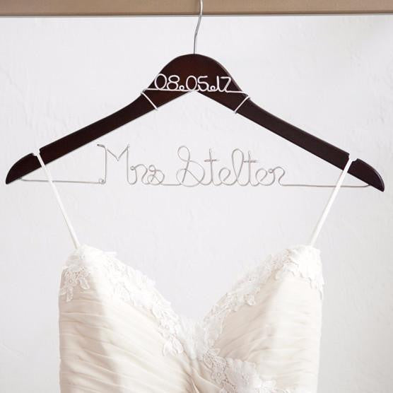 Dark wooden hanger with personalized date 08.05.17 in silver aluminum wire and base wire shaped into personalized name "Mrs Stelter" in silver. Holding a white wedding gown on a white background.