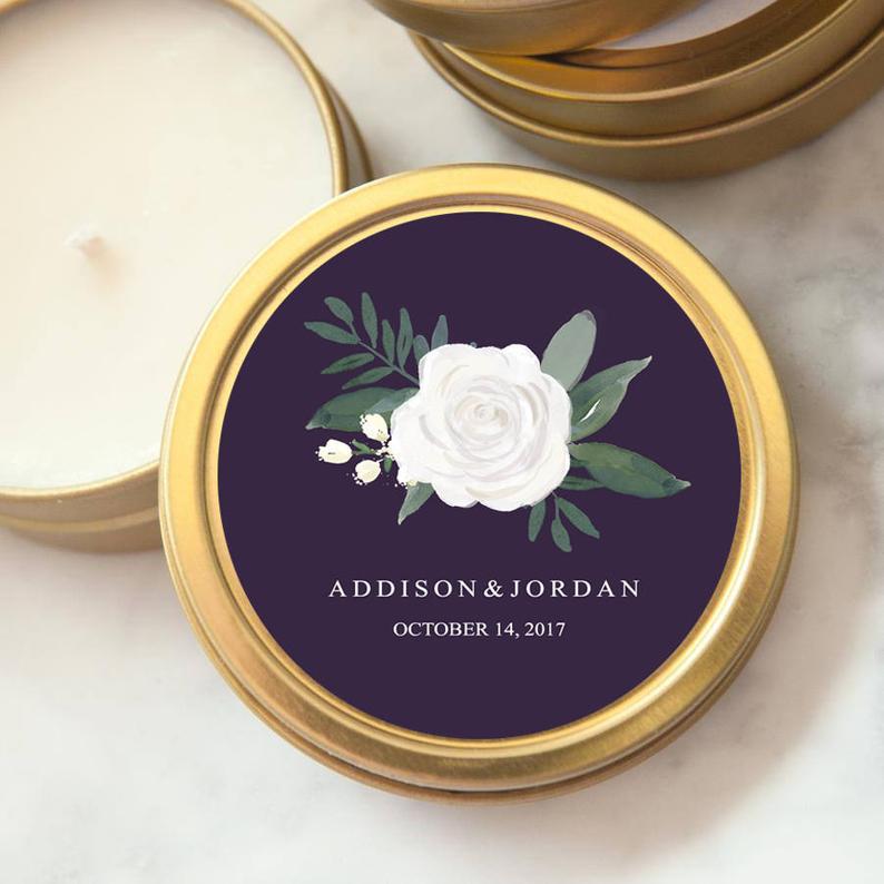 A detailed close-up image showcases a gold-rimmed candle tin adorned with a floral design, perfect for wedding or special event favors. The composition focuses on the candle tin's label, centered within a round frame. The label features a eggplant background with a delicate watercolor painting of a white rose, green leaves, and sprigs. Below the floral design, elegant typography reads "ADDISON & JORDAN" followed by "OCTOBER 14, 2017" in white lettering.