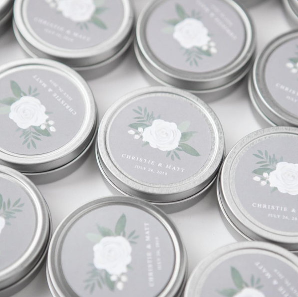  Image that showcases several silver-rimmed candle tins adorned with a floral design, perfect for wedding or special event favors. The composition focuses on the candle tin's label, centered within a round frame. The label features a grey background with a delicate watercolor painting of a white rose, green leaves, and sprigs. Below the floral design, elegant typography reads "Christie & Matt" followed by "July 26, 2019" in white lettering.