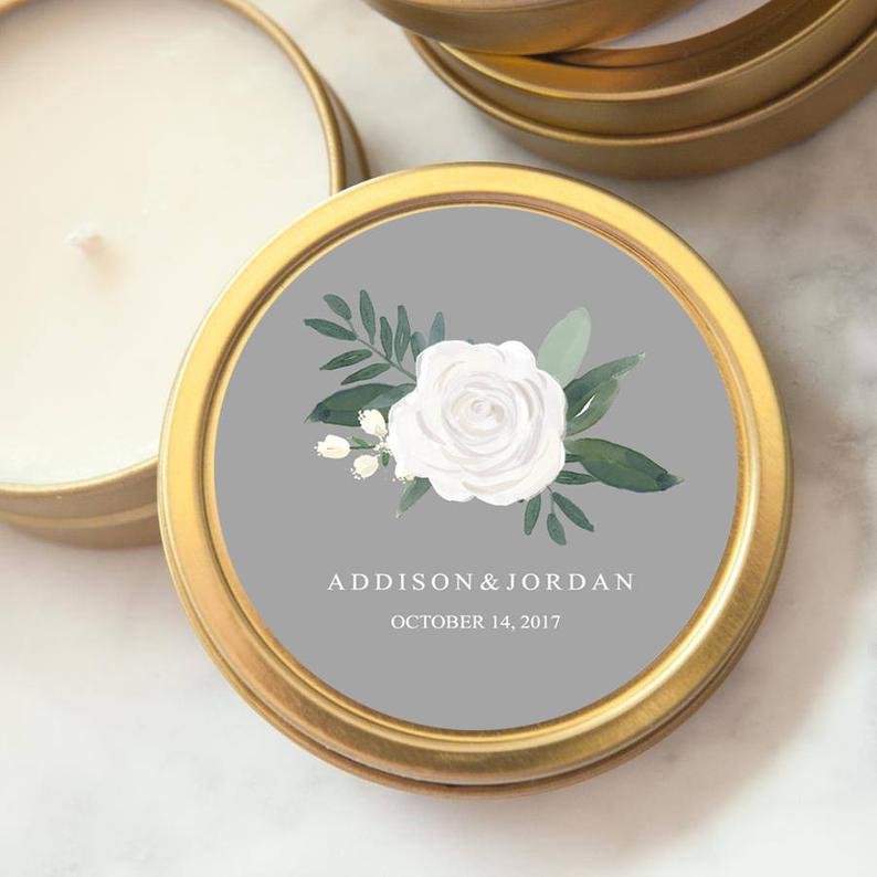A detailed close-up image showcases a gold-rimmed candle tin adorned with a floral design, perfect for wedding or special event favors. The composition focuses on the candle tin's label, centered within a round frame. The label features a grey background with a delicate watercolor painting of a white rose, green leaves, and sprigs. Below the floral design, elegant typography reads "ADDISON & JORDAN" followed by "OCTOBER 14, 2017" in white lettering.