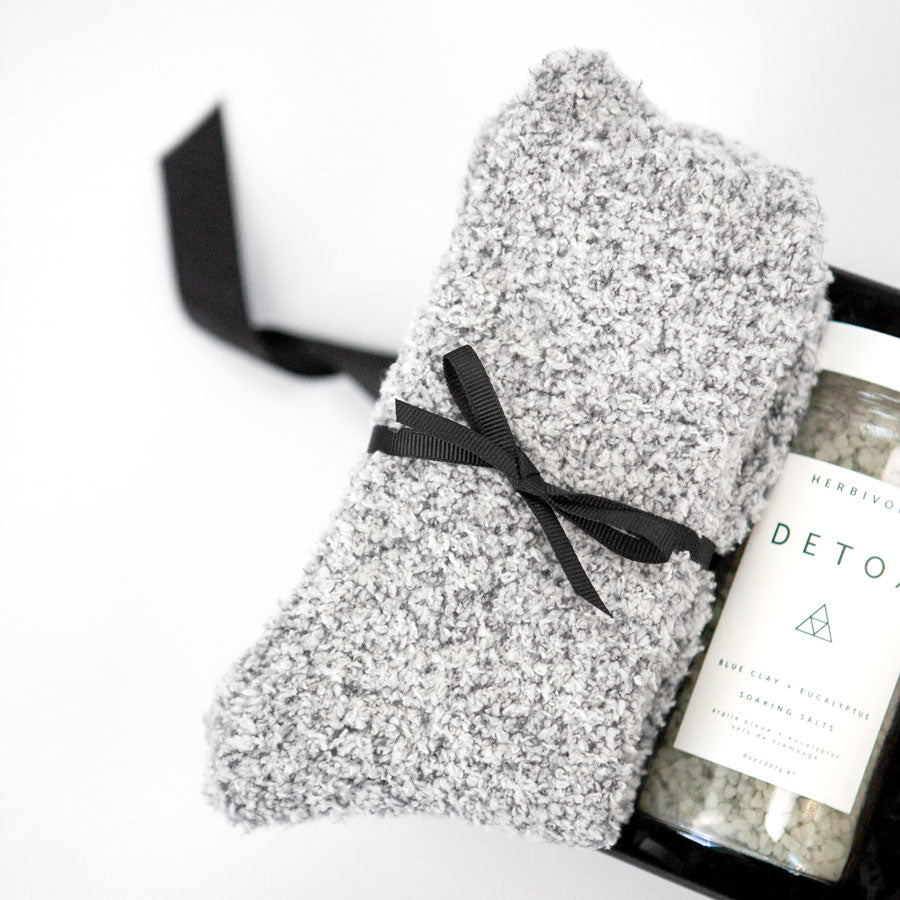 Gray 'foxblossom' socks with a black ribbon, 'herbivore' detox soaking salts in a glass jar, two 'breathe deeply' wipes in a white package, and black eucalyptus essential oil box and bottle, placed in a small black 'foxblossom' box with a black ribbon