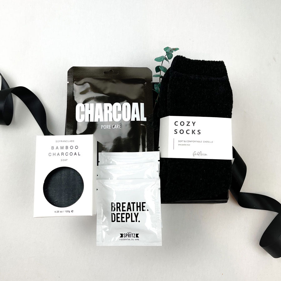 Black bamboo charcoal soap in a white box with viewing window, Lapcos charcoal pore care face mask, sprig of eucalyptus, black chenille unisex cozy socks and 3 white  packets of Spritz breathe deeply essential oil wipes. All arranged on a black gift box with a black satin ribbon. 