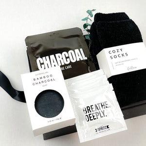 Black bamboo charcoal soap in a white box with viewing window, Lapcos charcoal pore care face mask, sprig of eucalyptus, black chenille unisex cozy socks and 3 white packets of Spritz breathe deeply essential oil wipes. All arranged on a black gift box with a black satin ribbon.