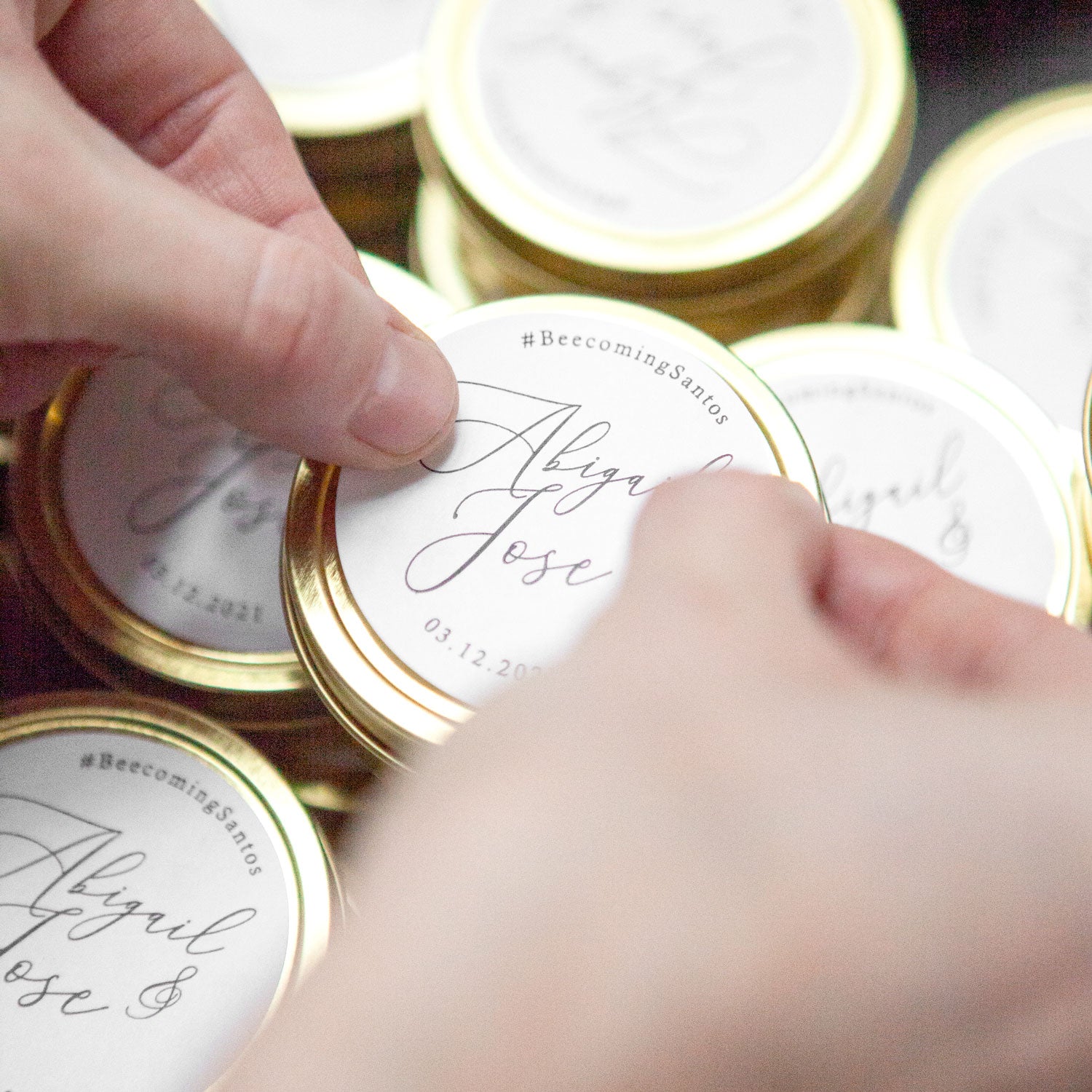 A close-up photograph displays a gold-colored candle tin featuring personalized details. a candle tin made of gold-colored metal, with a round shape and a visible seam. Inside the tin, there is a white candle. A circular label covers the top, displaying elegant calligraphy. The label features the names "Jenna & Mike," the hashtag "#becomingsantos"," and the date "03.12.2020."