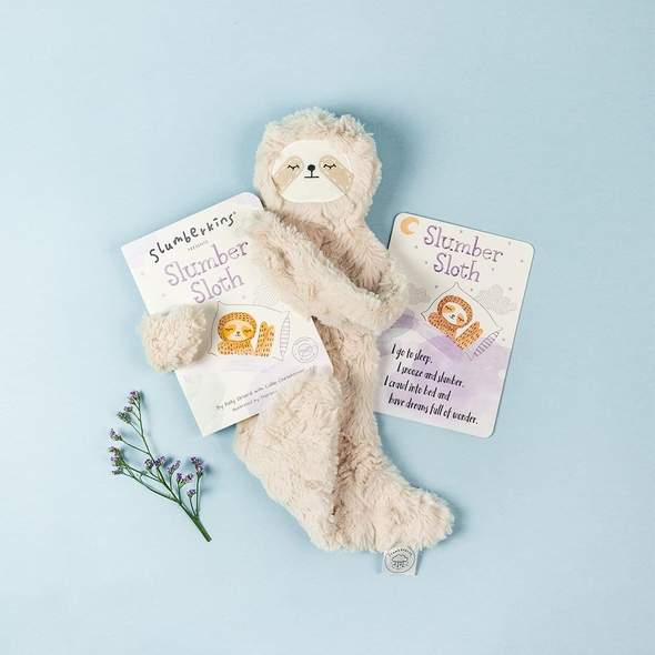 Slumberkins Slumber Sloth stuffed toy with Slumber Sloth hard book. Shown on a blue background with a floral accent. Key product in our Welcome Slumber gift box. 