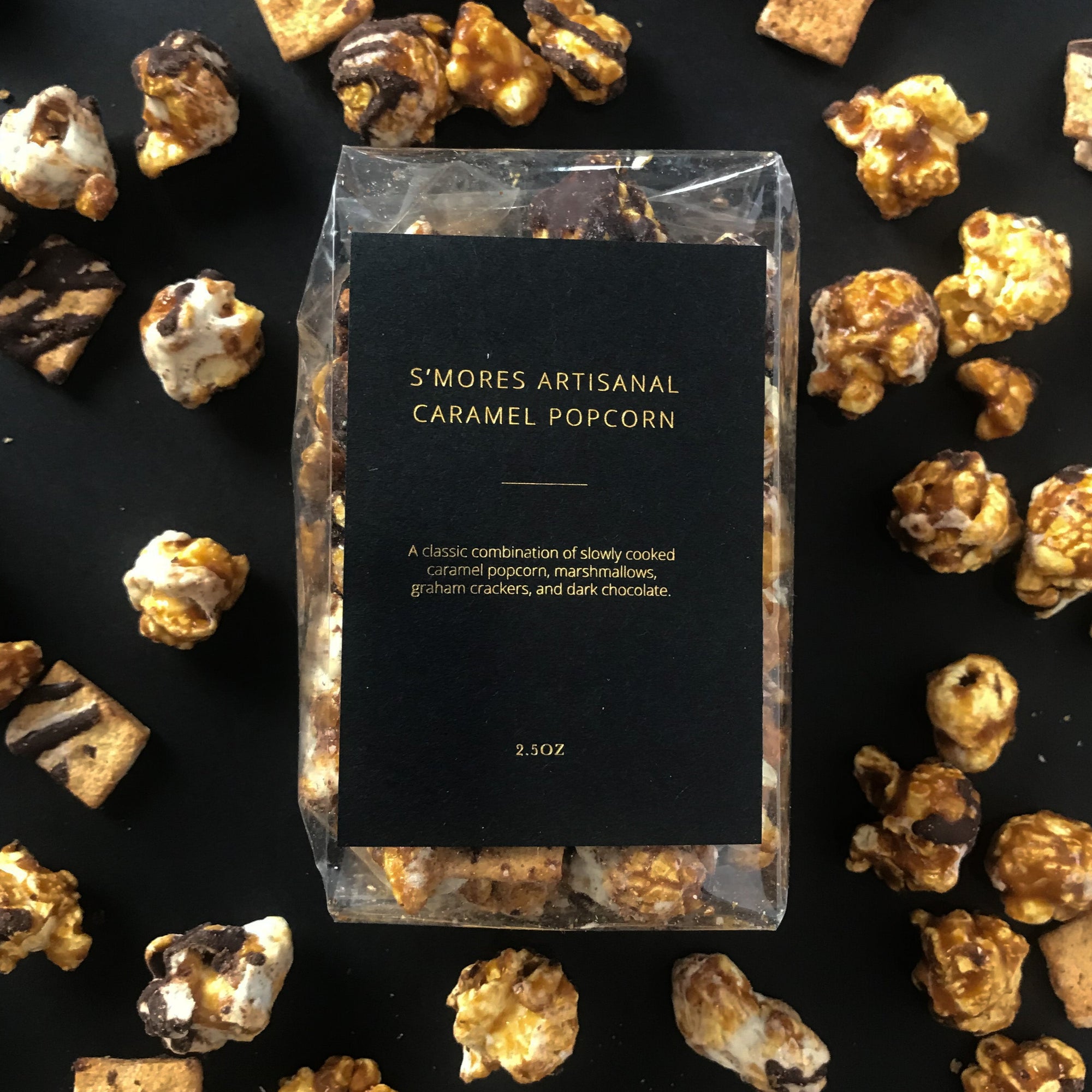 S'mores Artisanal Caramel Popcorn featured in its transparent bag with black label and gold lettering on a black background with popcorn around it. 