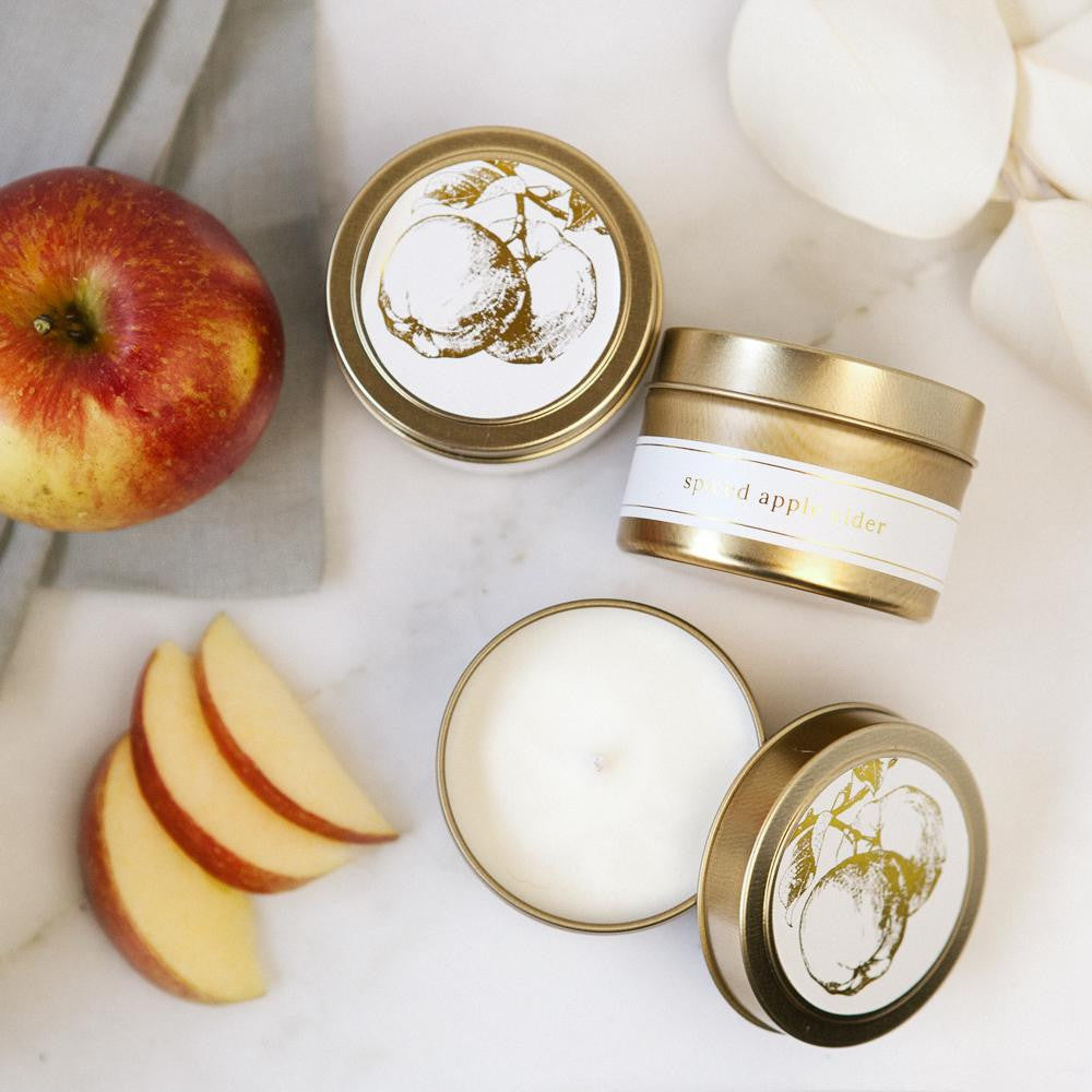Featuring three gold tin jar candles labeled “Spiced Apple Cider.” The candles are displayed in different positions: one viewed from the top with its lid on, one placed sideways to reveal the side label, and one opened to show the smoot white wax inside. Each tin has a clean white label with elegant gold lettering and a delicate gold illustration of an apple tree on the lid.  Set against a soft background with scattered red apple slices and white rose petals. A folded grey cloth in the corner.