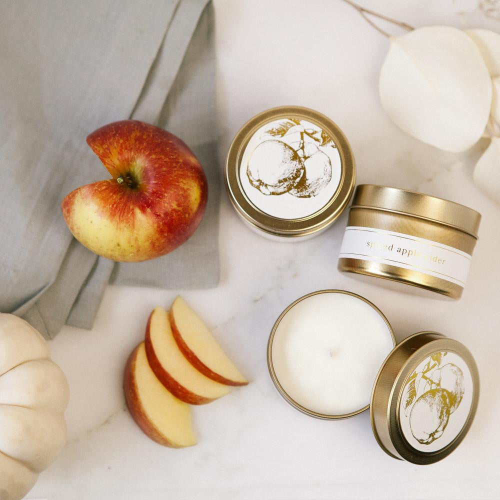 Featuring three gold tin jar candles labeled “Spiced Apple Cider.” The candles are displayed in different positions: one viewed from the top with its lid on, one placed sideways to reveal the side label, and one opened to show the smoot white wax inside. Each tin has a clean white label with elegant gold lettering and a delicate gold illustration of an apple tree on the lid.  Set against a soft background with scattered red apple slices and white rose petals. A folded grey cloth in the corner.