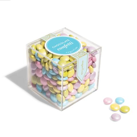 Sugarfina transparent plastic cube filled with colorful candy-coated chocolate pieces with blue label, white lettering, and gold details.