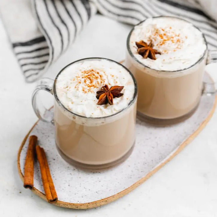 Two mugs of warm, tea drinks sit on a white platter, topped with whipped cream and star anise.Two cinnamon sticks rest on the platter next to the mugs.  