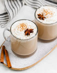 Two mugs of warm, tea drinks sit on a white platter, topped with whipped cream and star anise.Two cinnamon sticks rest on the platter next to the mugs.  