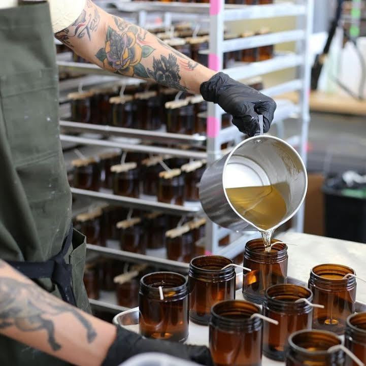 A teakwood and tobacco candle is shown being poured into its jar by a man with black gloves in the production headquarters. 
