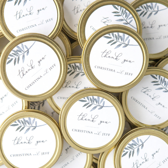 Thank you custom wedding favor candle featured image presents a collection of small, gold-rimmed tins, each with a label reading "thank you" along with the names "CHRISTINA and JEFF,"  watercolor-style depiction of a leafy branch in shades of grey-blue is placed near the top. The tins themselves have a classic, metallic gold finish.