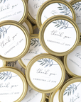 Thank you custom wedding favor candle featured image presents a collection of small, gold-rimmed tins, each with a label reading "thank you" along with the names "CHRISTINA and JEFF,"  watercolor-style depiction of a leafy branch in shades of grey-blue is placed near the top. The tins themselves have a classic, metallic gold finish.