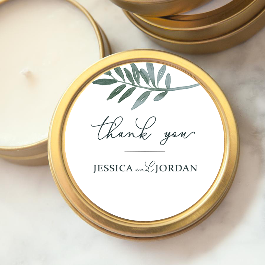 Close-up image of the thank-you custom wedding favor candle in its gold-rimmed tin, each with a label reading "Thank You" along with the names "Jessica and Jordan." A watercolor-style depiction of a leafy branch in shades of grey-blue is placed near the top.  The lid opened, showing the white candle on a marble-like background. 