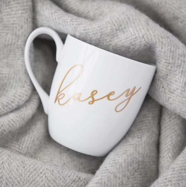 White personalized mug in gold, pictured with a grey scarf in the background. 