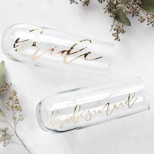 Two personalized champagne flutes, read "bride" and "bridesmaid" in mirrored gold script.