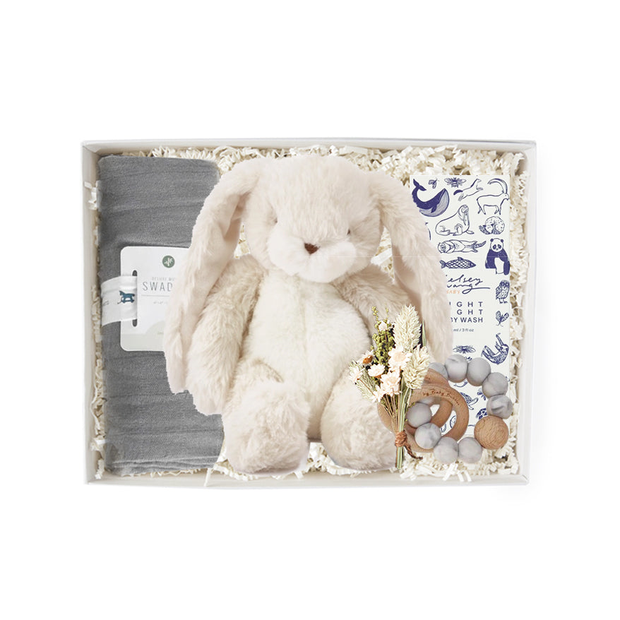 A gray swaddle blanket, a white stuffed bunny, floral bundle, wooden baby rattle, and animal graphic printed box of 'night night body wash' in a gray 'foxblossom' box with ivory paper shread.